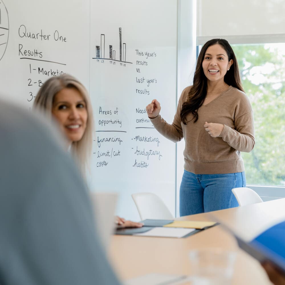 A woman stands next to a white board in a conference room while talking with her colleagues.