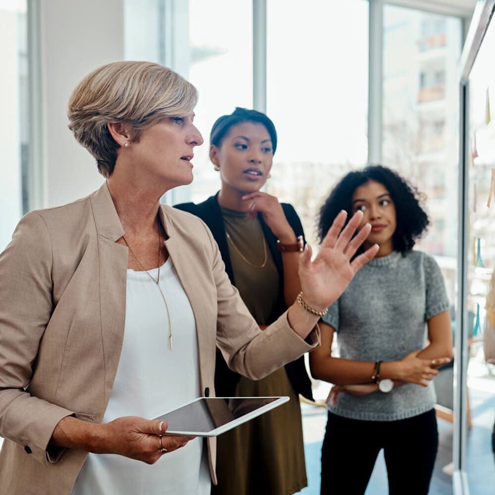 A group of three woman stand in front of a whiteboard with sticky notes, while one is talking and pointing.