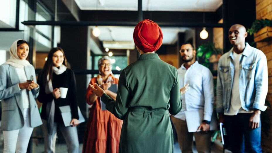 A person with a head wrap talks in front of a group of people while everyone is standing.