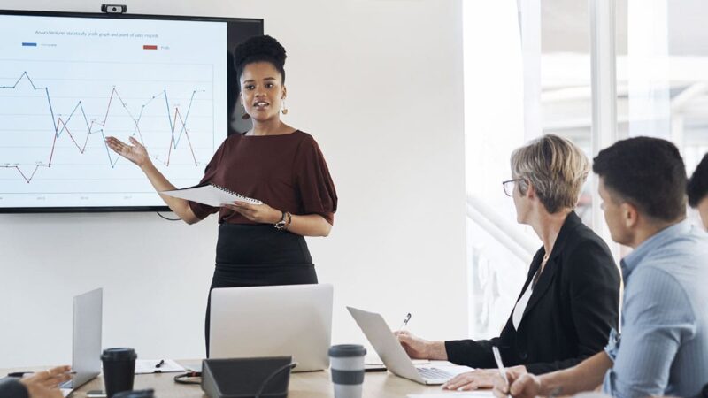A Black woman leads a presentation in front of a conference room of people.
