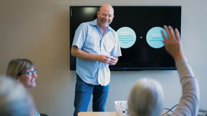A man leads a presentation in a conference room, while an attendee raises their hand for a question.