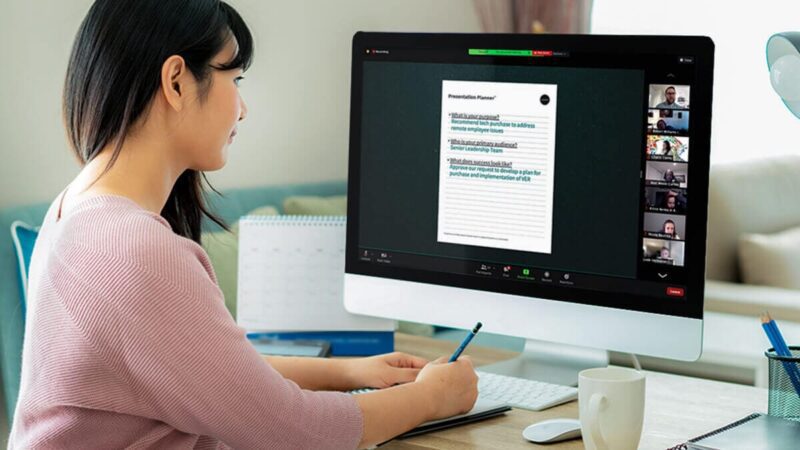 A woman takes notes while watching a virtual presentation on her computer.