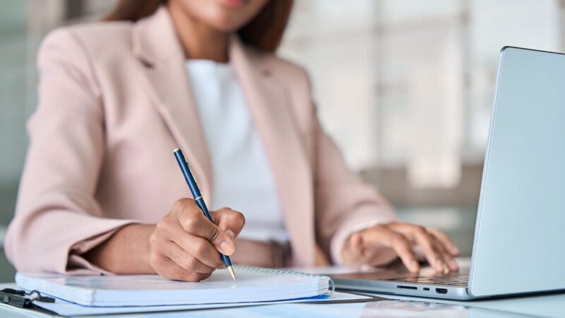 A woman on a laptop writes notes on a pad of paper for the Virtual Presentation Checklist.