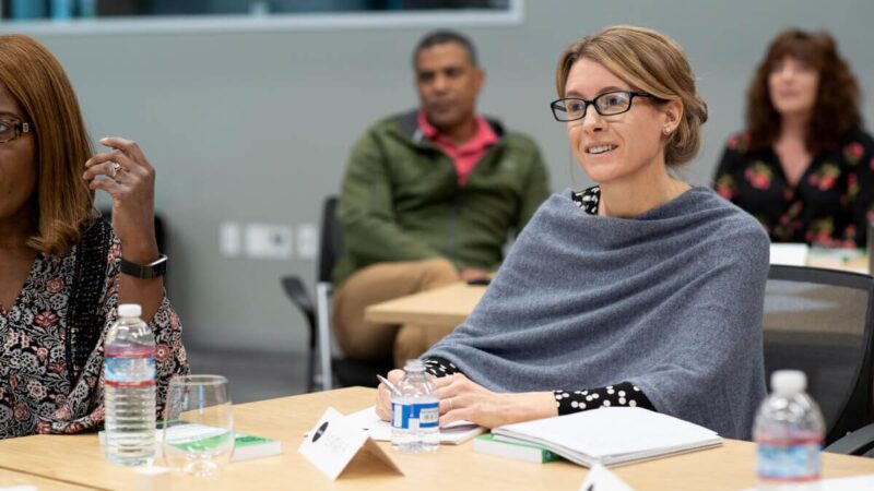A woman watches a presentation while taking notes in her notebook, in a classroom with other students.