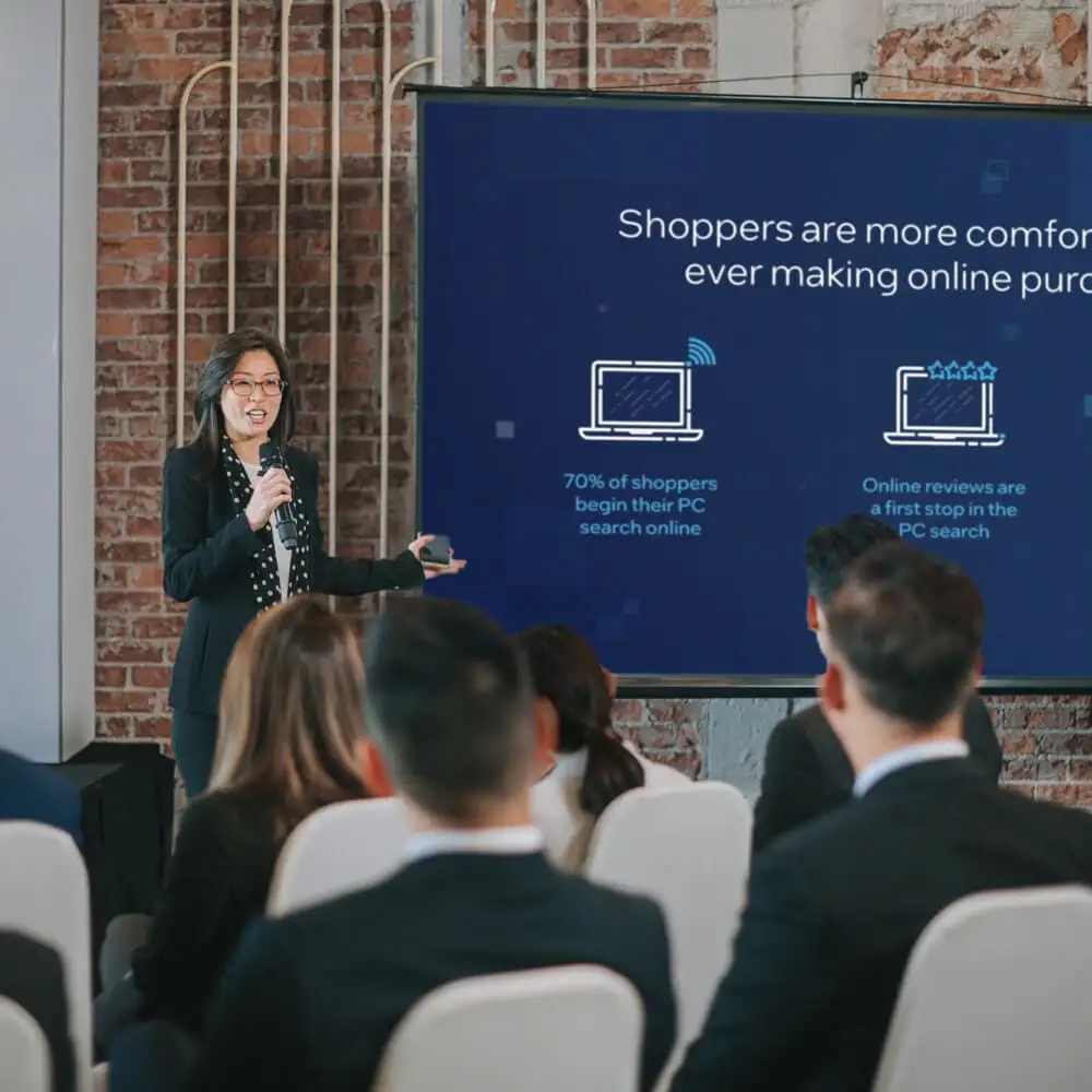 An Asian woman leads a presentation in front of a group of people at a conference.