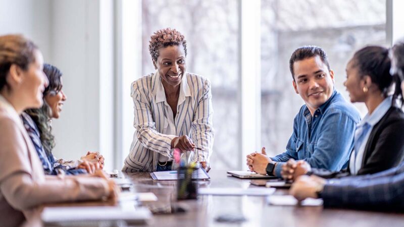 Image of co-workers in a meeting gathered around a conference table, with their team leader standing at the head of the table. Each person is practicing effective executive presence by looking confident, composed, and ready to meet the needs of their audience when it's their turn to speak.