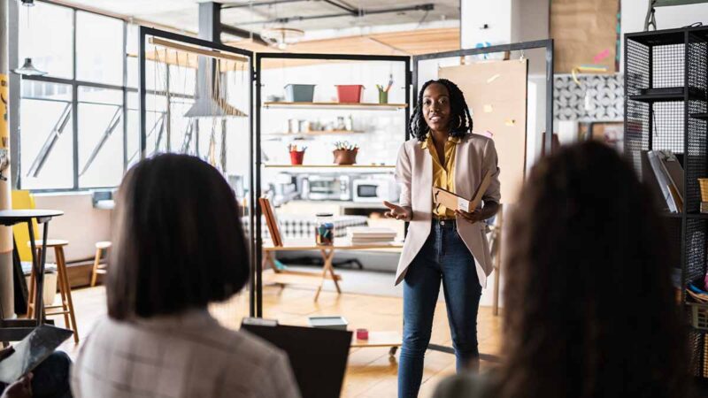 A professional standing in front of a group to offer their unique perspective during a team meeting. This demonstrates how understanding the value of one’s contributions and sharing them confidently is essential to building executive presence.
