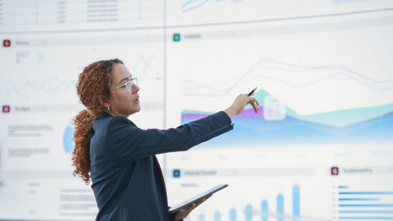 A professional is standing in front of a line chart and pointing toward a specific peak. This illustrates how Duarte’s Skills Roadmap for Data Storytelling can help turn data explorers into data leaders through structured skill-building.