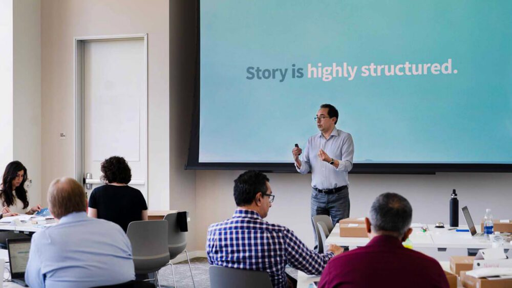 A top-rated Duarte facilitator conducting an in-person DataStory training workshop. The facilitator is standing in front of screen featuring the words “Story is highly structured” while a group of workshop attendees are facing front with rapt attention.