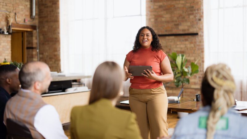 A professional is standing at the front of the room, addressing a seated audience. They're smiling, their posture is relaxed, and they're communicating under pressure with confidence.