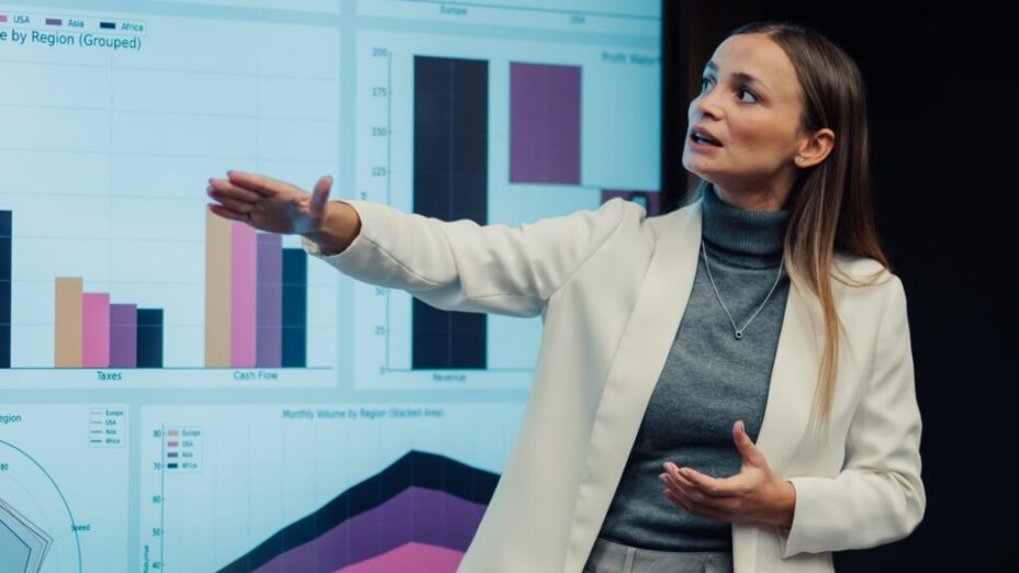 A business professional is giving a presentation and looking over their shoulder at the data displayed on screen behind them. They have an uncertain expression on their face as if they're not confident in how they're presenting data insights.