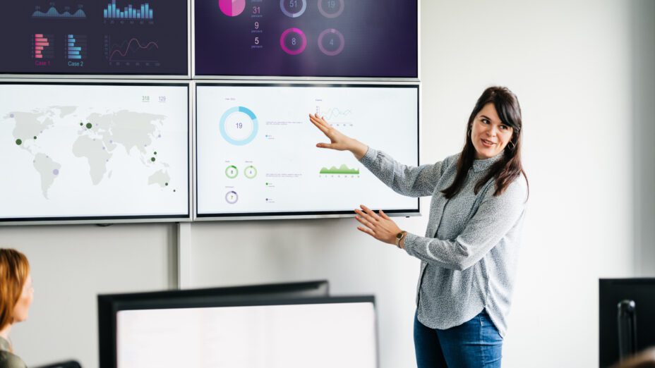 A presenter is standing in front of a digital screen with images, graphs, and charts displayed. Their co-workers are seated in front of them taking in the visuals.