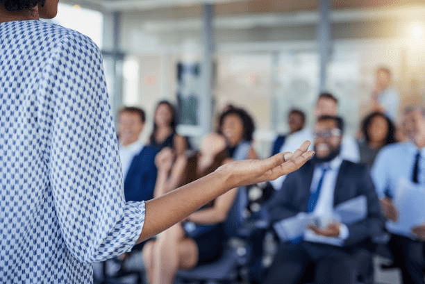 Person standing addressing a group of professionals in business attaire.