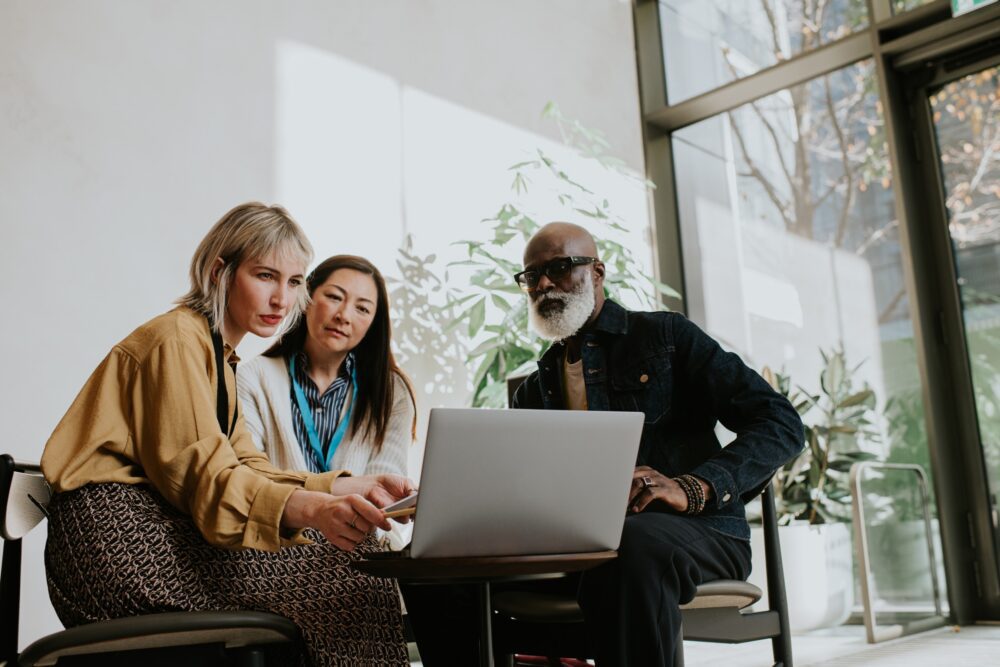 Three people reviewing a laptop together by a window with plants in the background.