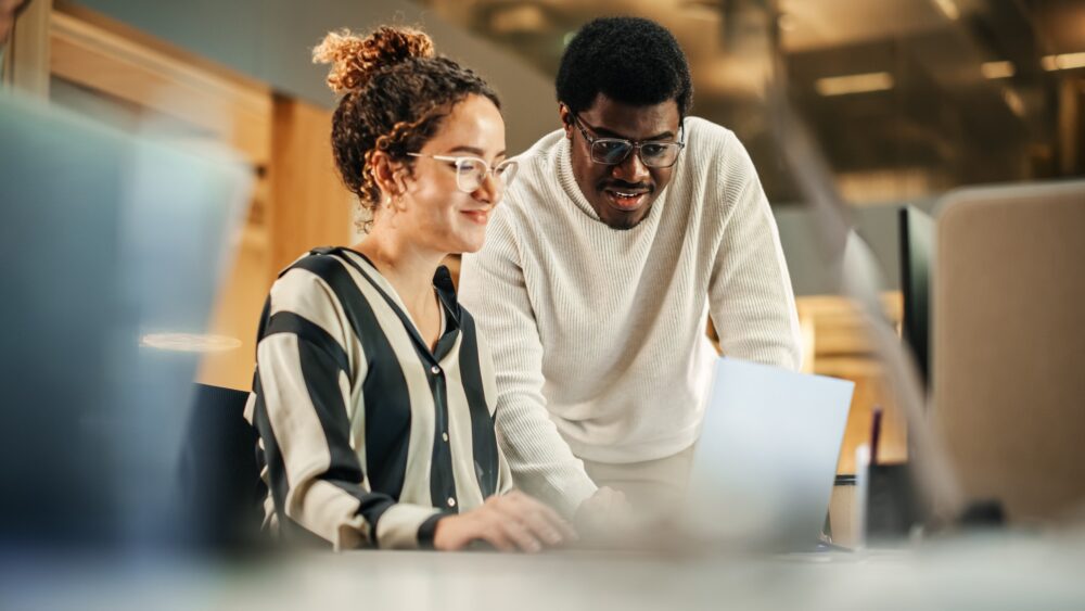 Two colleagues smiling while looking at a laptop screen in a modern office setting.