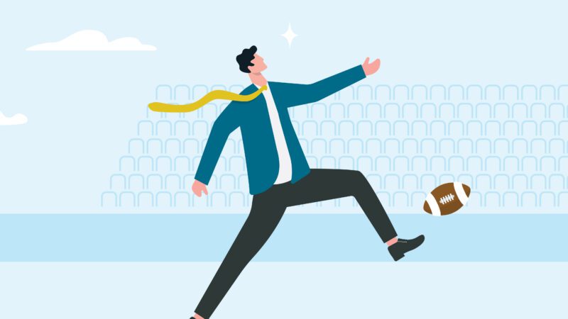 person in business attire kicking an american football as their tie blows in the wind.