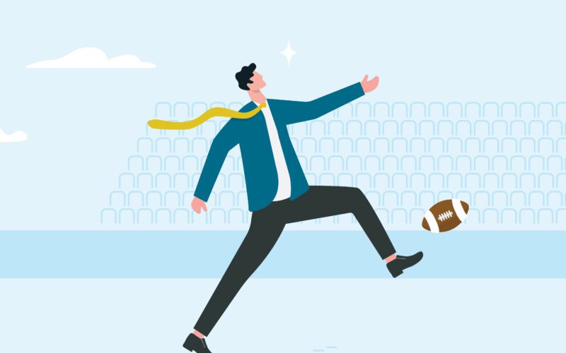 person in business attire kicking an american football as their tie blows in the wind.