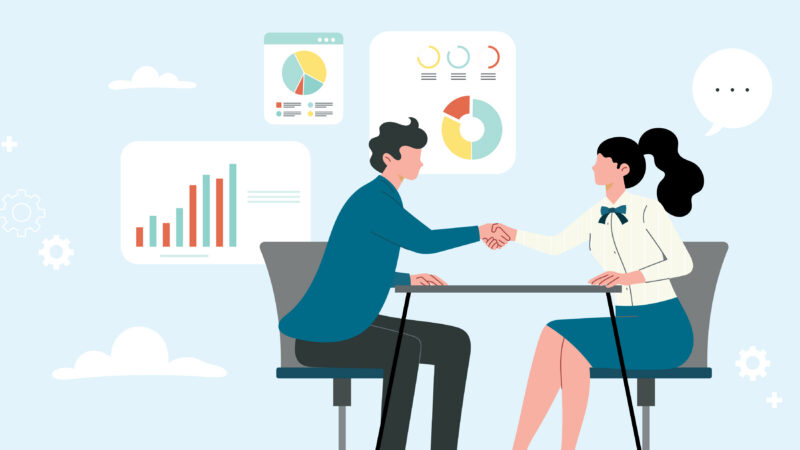A seller and a buyer shake hands across a table in front of sales performance charts after a successful pitch.